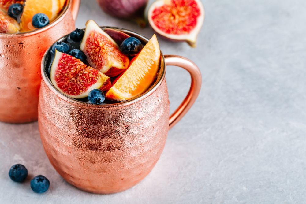 Copper mugs filled with a fruit cocktail featuring figs, oranges, and blueberries, set on a gray surface.