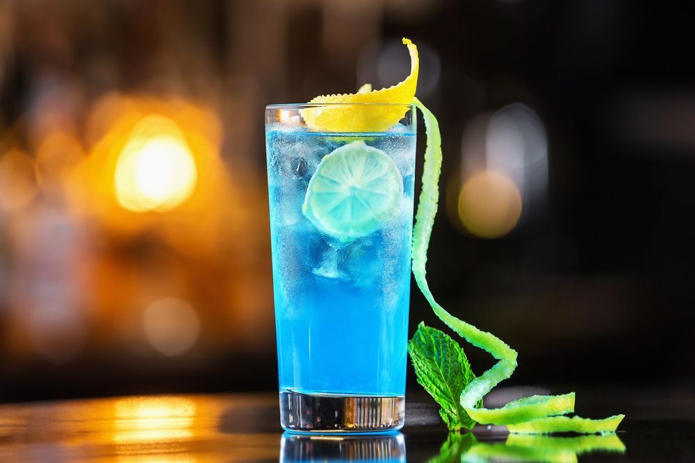 Blue cocktail in a tall glass garnished with lime and lemon twist, on a bar.