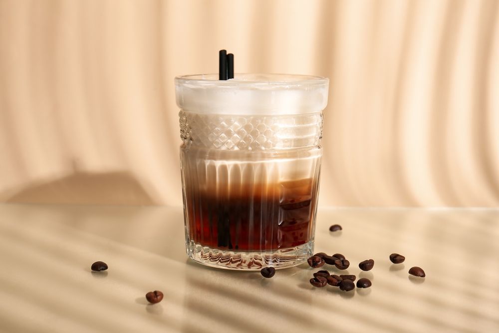 Cocktail in a patterned glass, layered with dark liquid, frothy white top, two black straws, coffee beans scattered around.