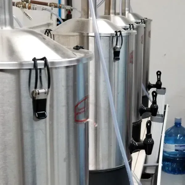 Four large stainless steel tanks with lids and spigots in a distillery. One blue water jug is in the background.