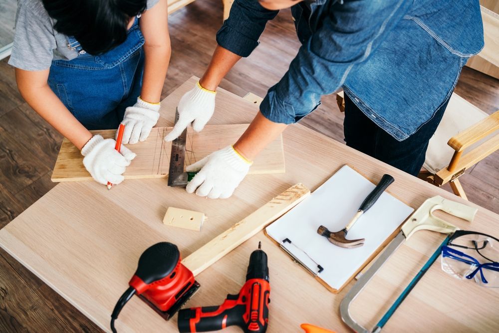 Two people using tools on wood, woodworking, indoor setting, wearing gloves.
