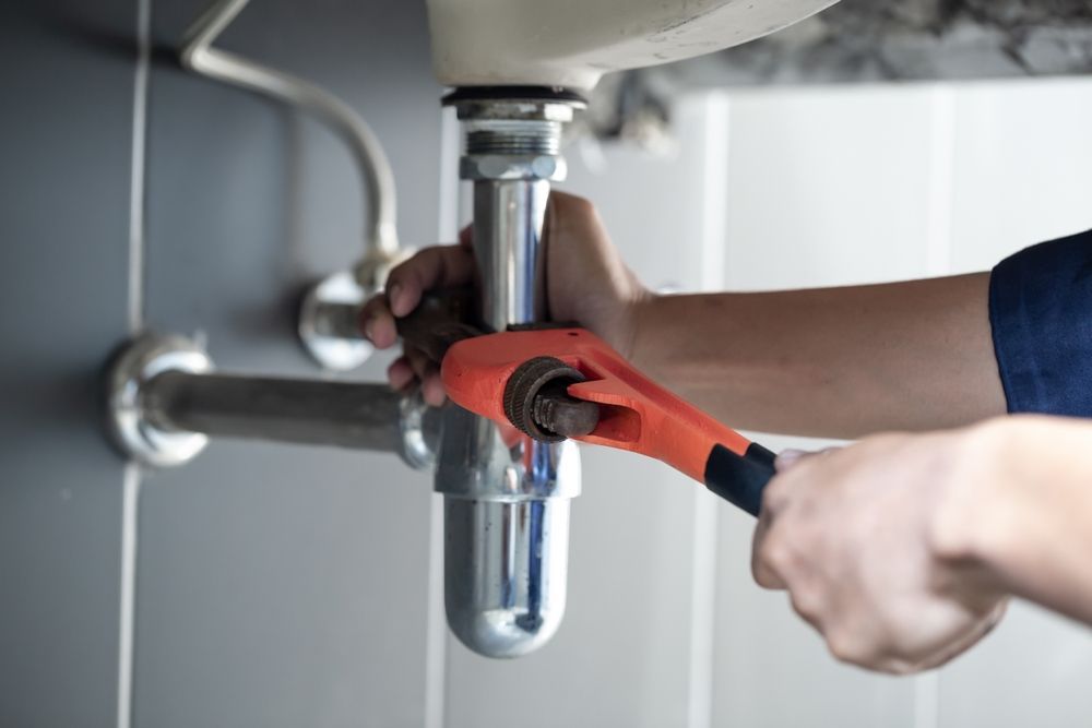 Plumber using a wrench to work on a sink's drain pipe, hands visible, interior setting.