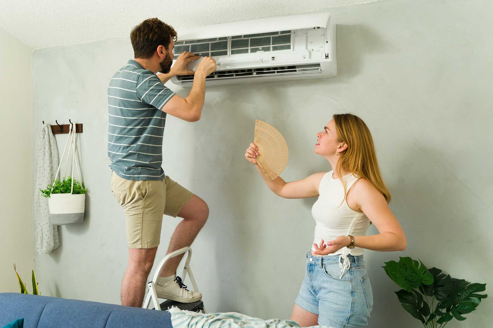 A man standing on a ladder trying to repair an AC unit while a woman fans herself.
