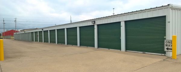 A row of storage units with green doors are lined up in a parking lot.