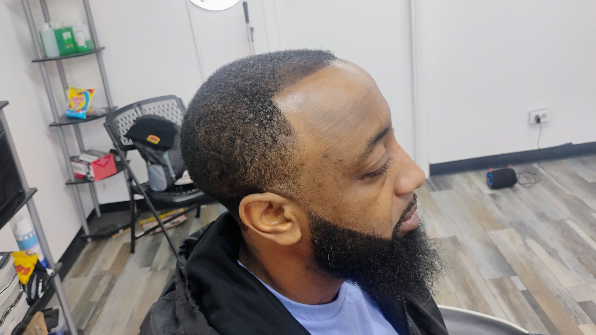 A side view of a person with a short, faded haircut and a dark, groomed beard, seated in a barbershop.