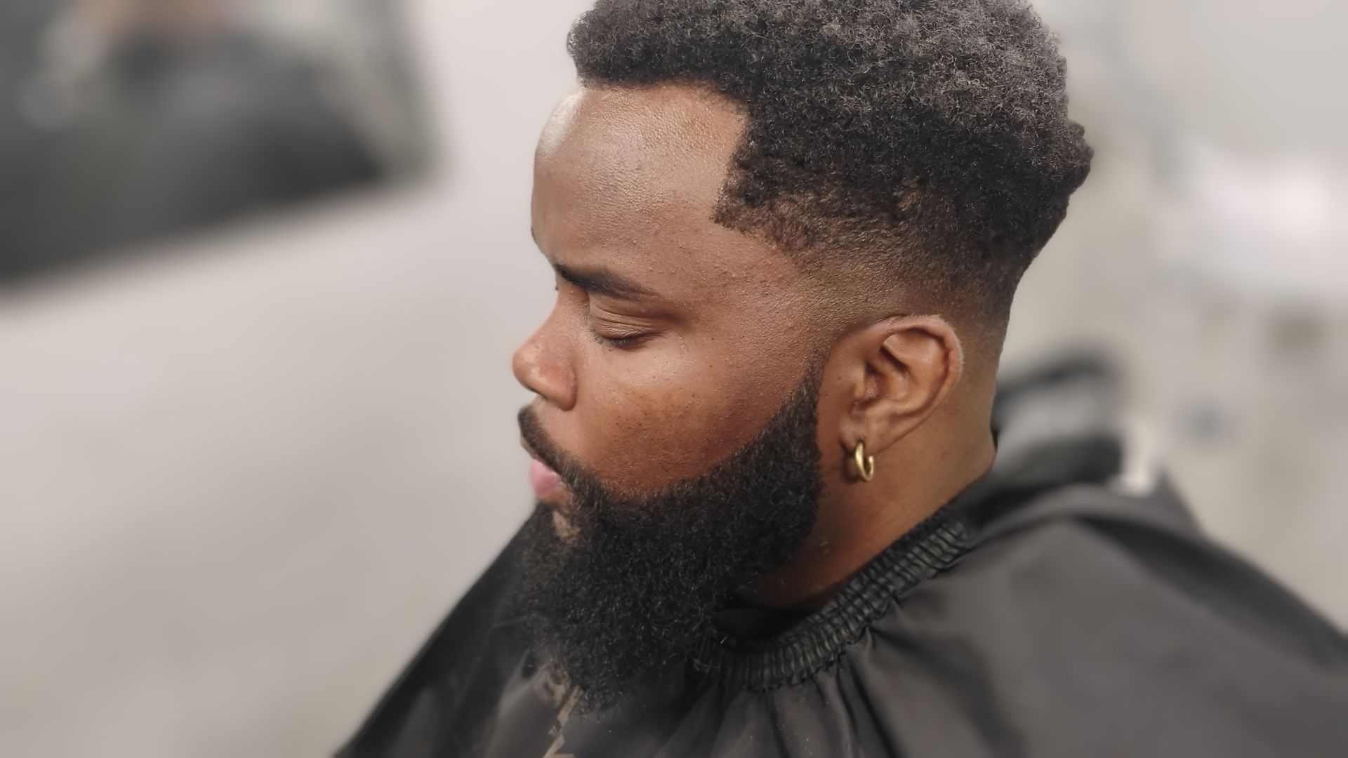 A profile view of a person with a precision-faded haircut, a styled beard, and a gold hoop earring in a salon setting.