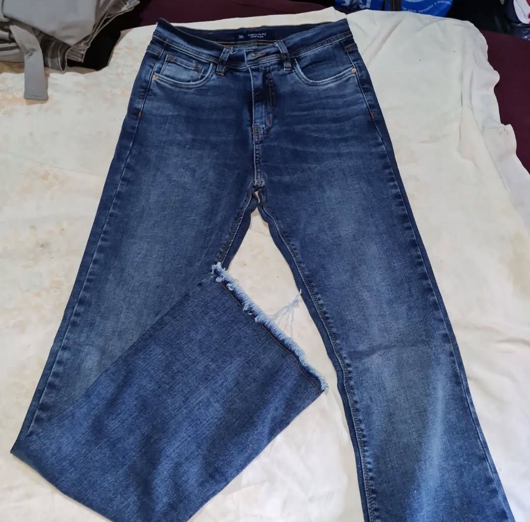 Vaqueros de mezclilla azul, de cintura alta, con dobladillo sin rematar y lavado descolorido, sobre una superficie blanca.