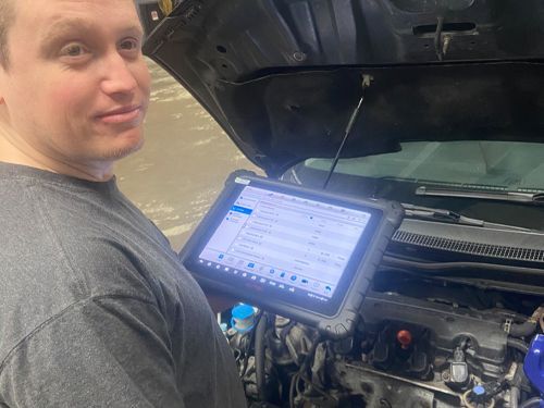 A man is standing in front of a car with the hood open and using a tablet.