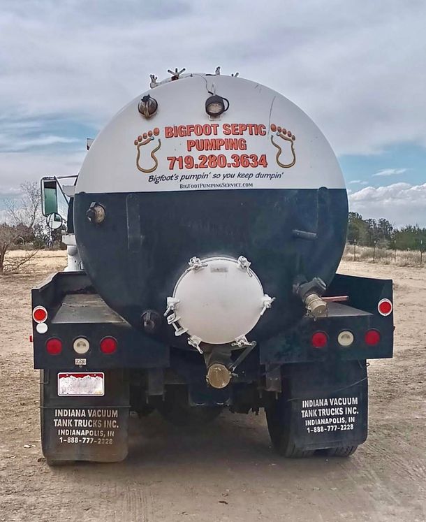 Man connecting green hose to septic tank truck, brown building in background.
