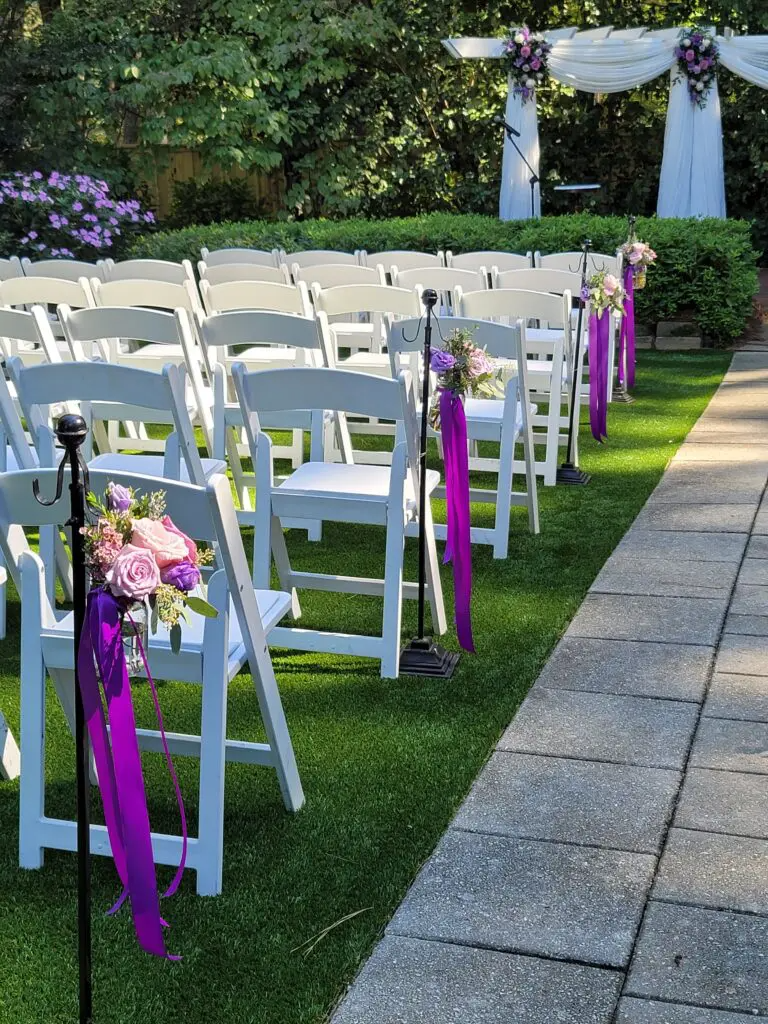 A row of white folding chairs decorated with purple ribbons and flowers