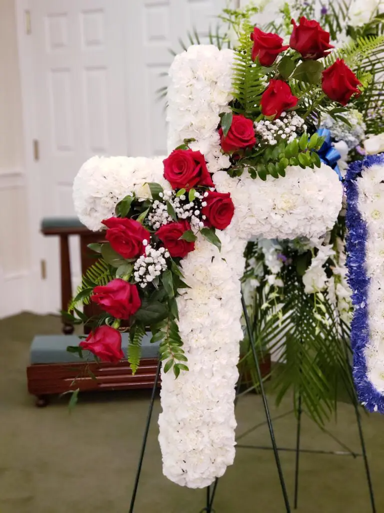 A white cross is decorated with red roses and baby 's breath.