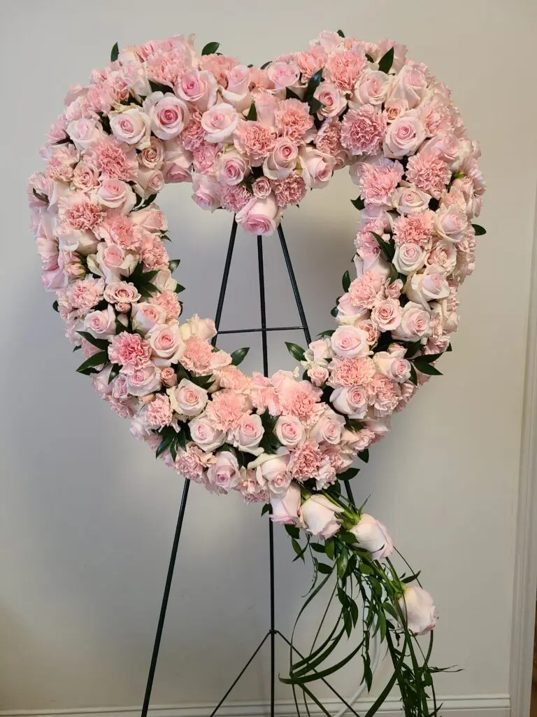 A heart shaped wreath made of pink roses and carnations on a stand.
