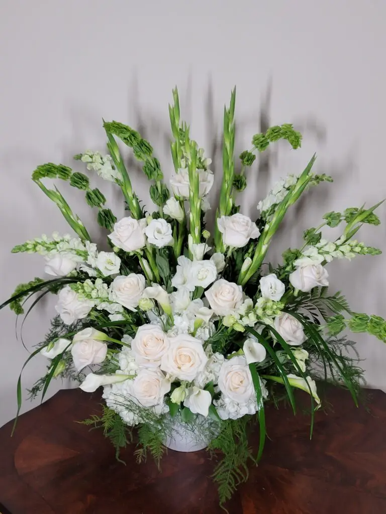 A vase filled with white flowers and greenery is sitting on a wooden table.