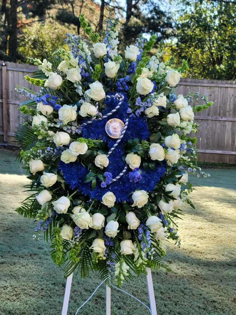 A wreath of blue and white flowers is sitting on a stand in front of a wooden fence.