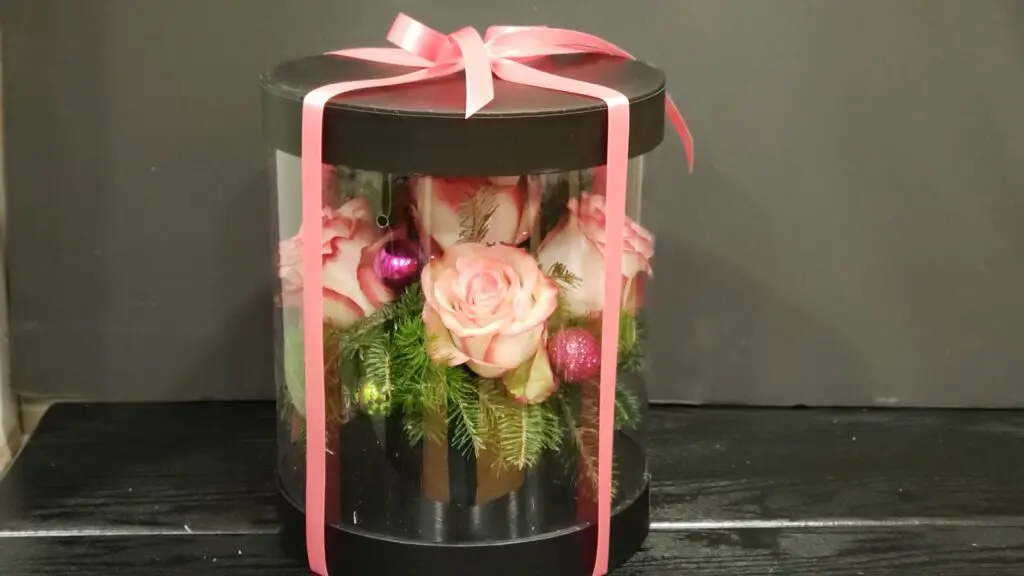 A clear box filled with pink roses and pink christmas ornaments.
