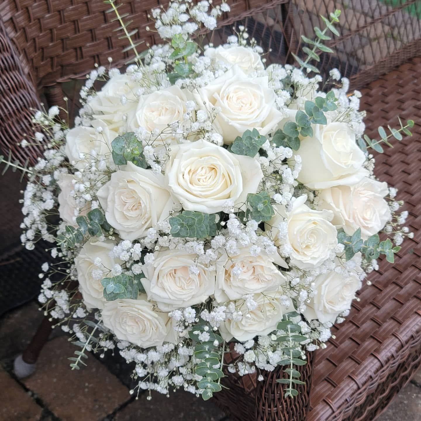 A bouquet of white roses and baby 's breath is sitting on a wicker chair.