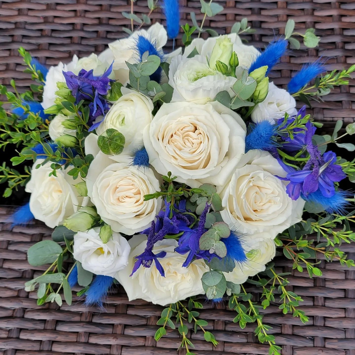 A bouquet of white roses and blue flowers is sitting on a wicker table.