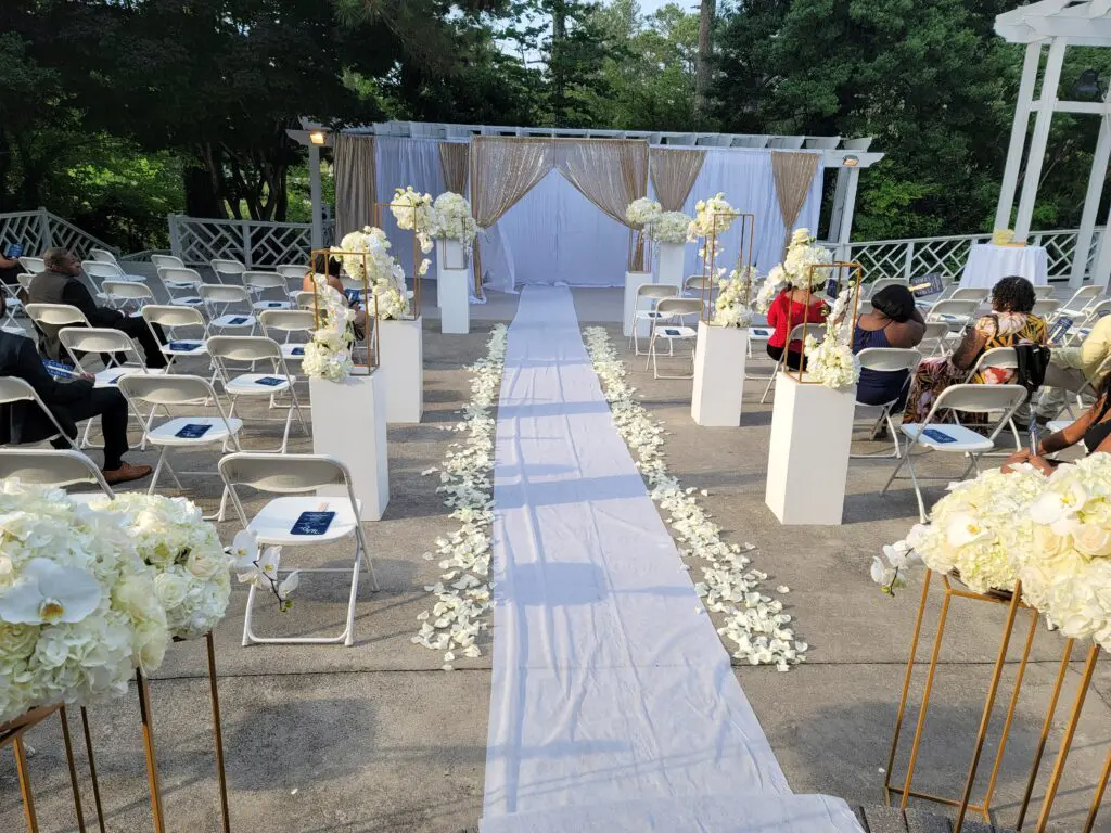 A wedding ceremony is being held in a park with a white aisle lined with white flowers.
