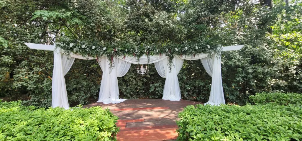 A white archway with white curtains is surrounded by trees and bushes.
