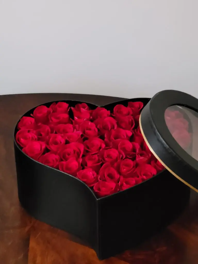 A heart shaped box filled with red roses is sitting on a wooden table.