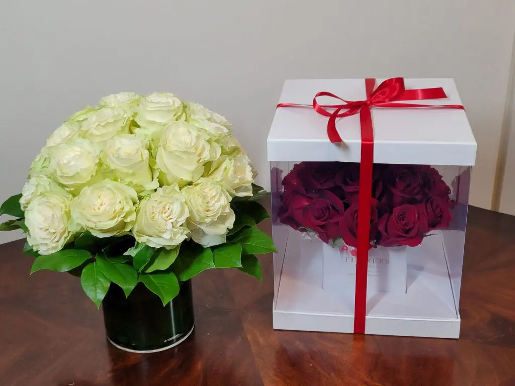 A vase of white roses next to a box of red roses on a table.