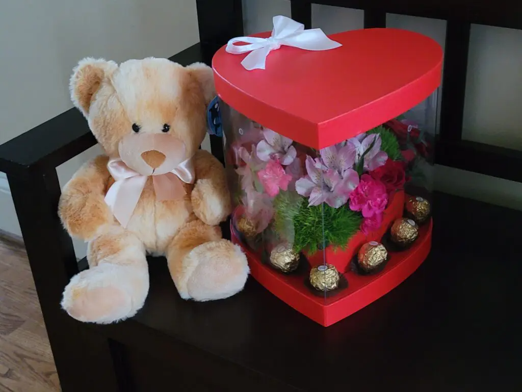 A teddy bear sits next to a heart shaped box filled with flowers and chocolates