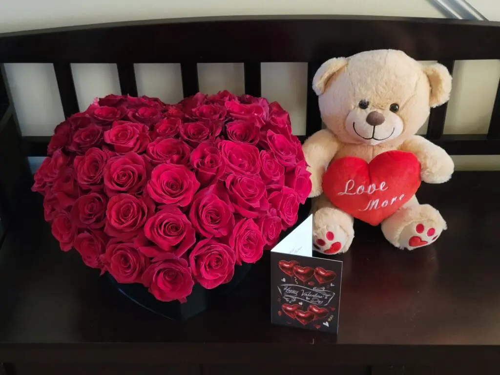 A teddy bear holding a heart next to a heart shaped bouquet of red roses