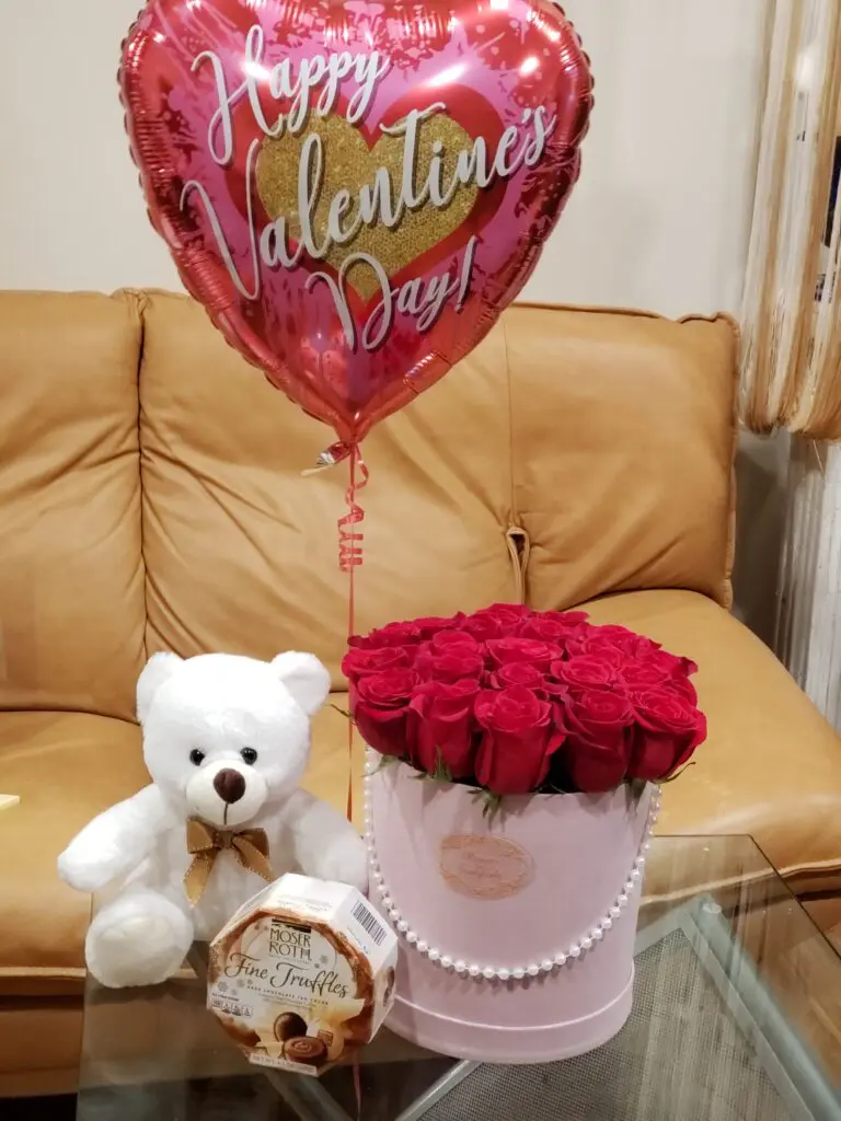 A valentine 's day balloon is sitting on a table next to a teddy bear and a box of red roses.