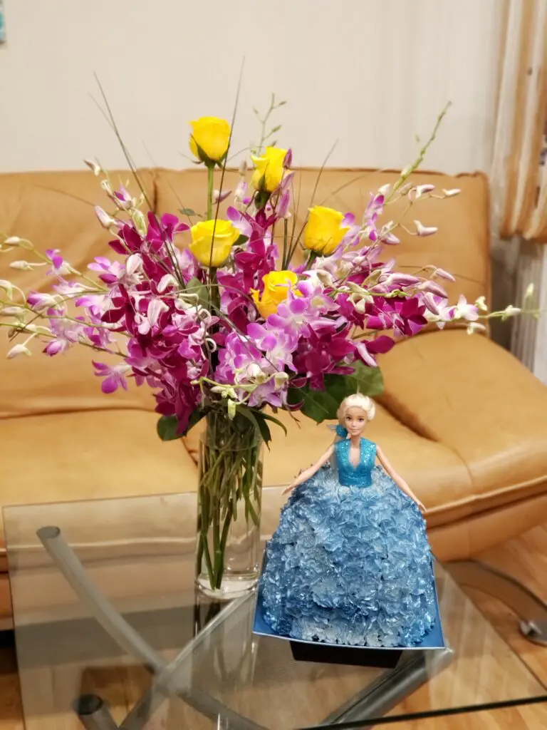 A doll in a blue dress is sitting on a table next to a vase of flowers