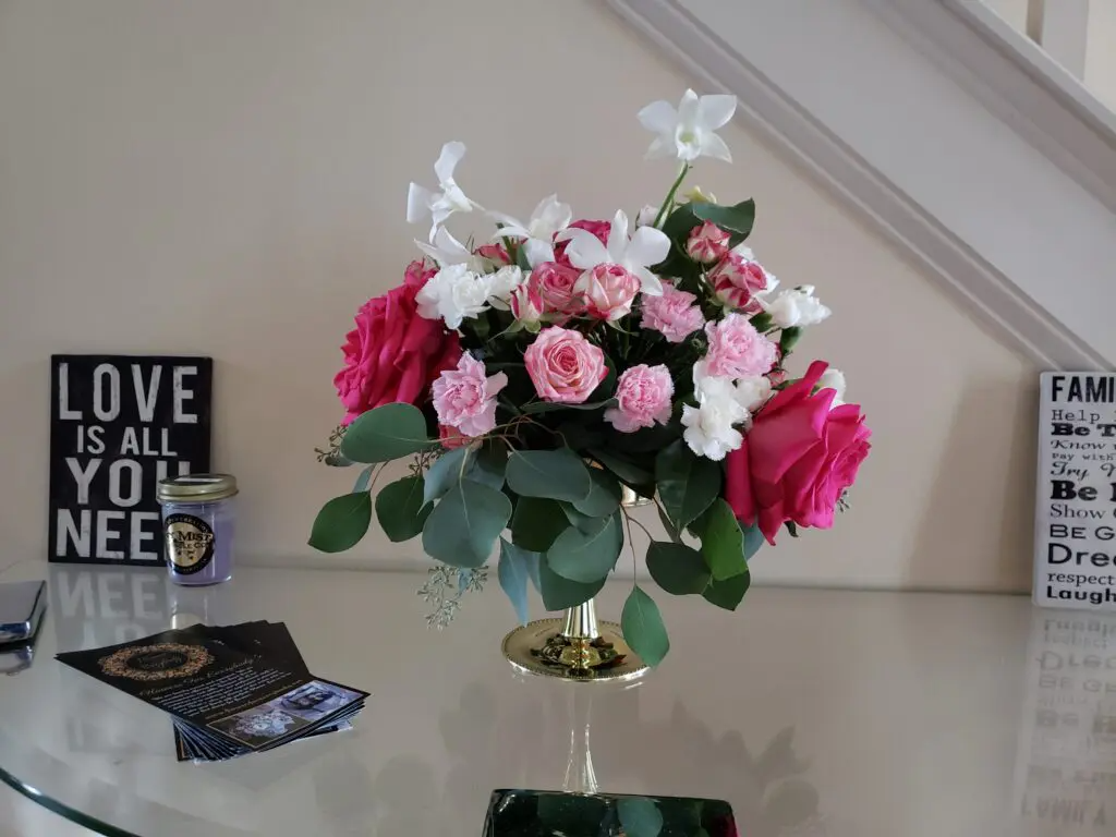 A vase filled with pink and white flowers is on a table next to a sign that says love is all you need.