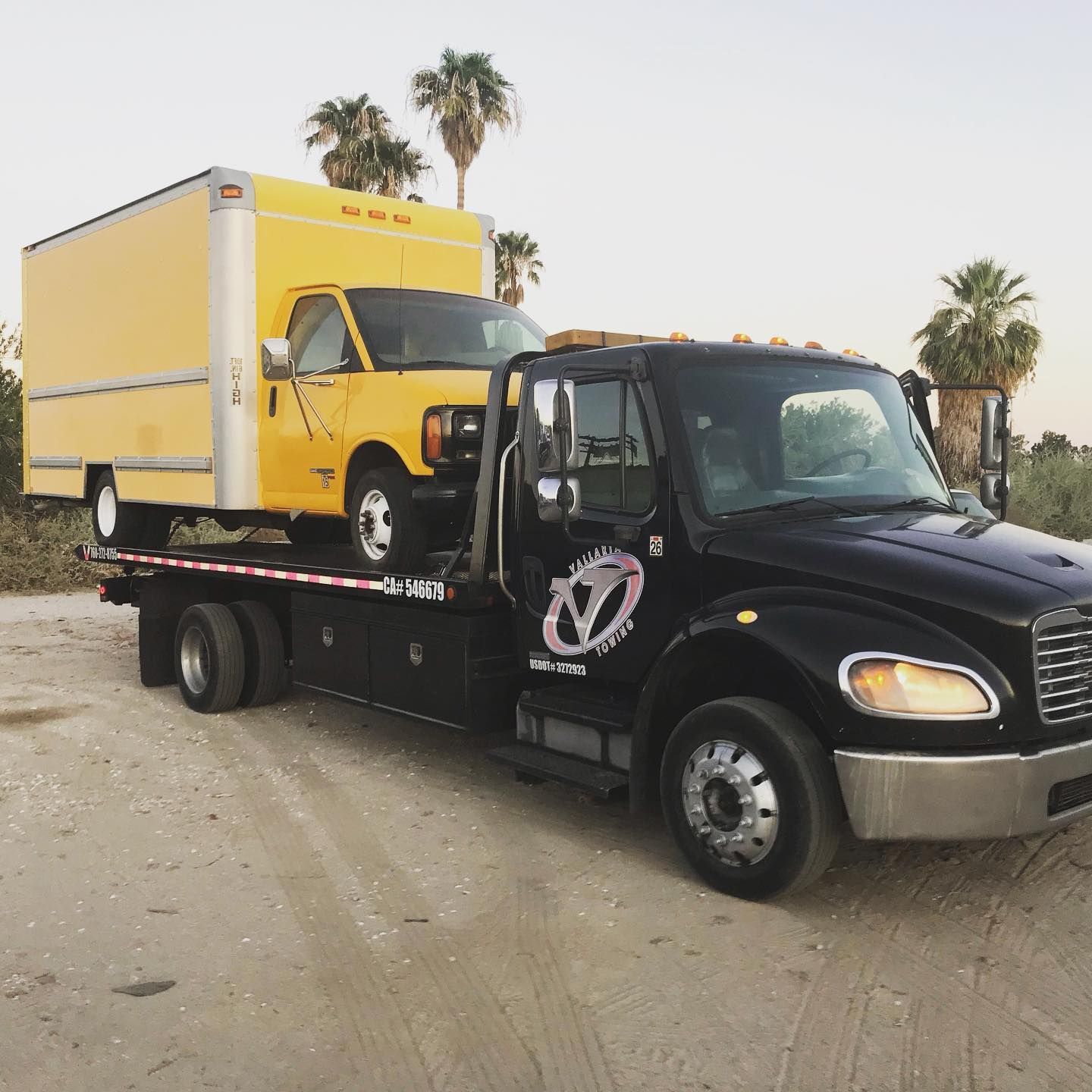 Black tow truck carrying a yellow box truck in a desert setting; palm trees in the background.