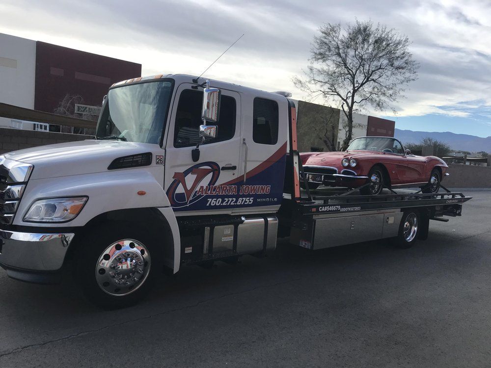 Tow truck with a red convertible on its flatbed. White truck, blue and red logo, desert setting.