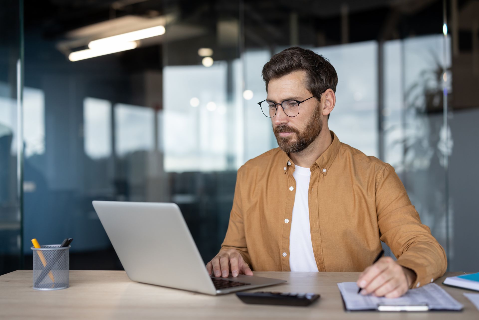 Ein Mann mit Brille arbeitet an einem Laptop an einem Schreibtisch in einem Büro mit Glaswänden;