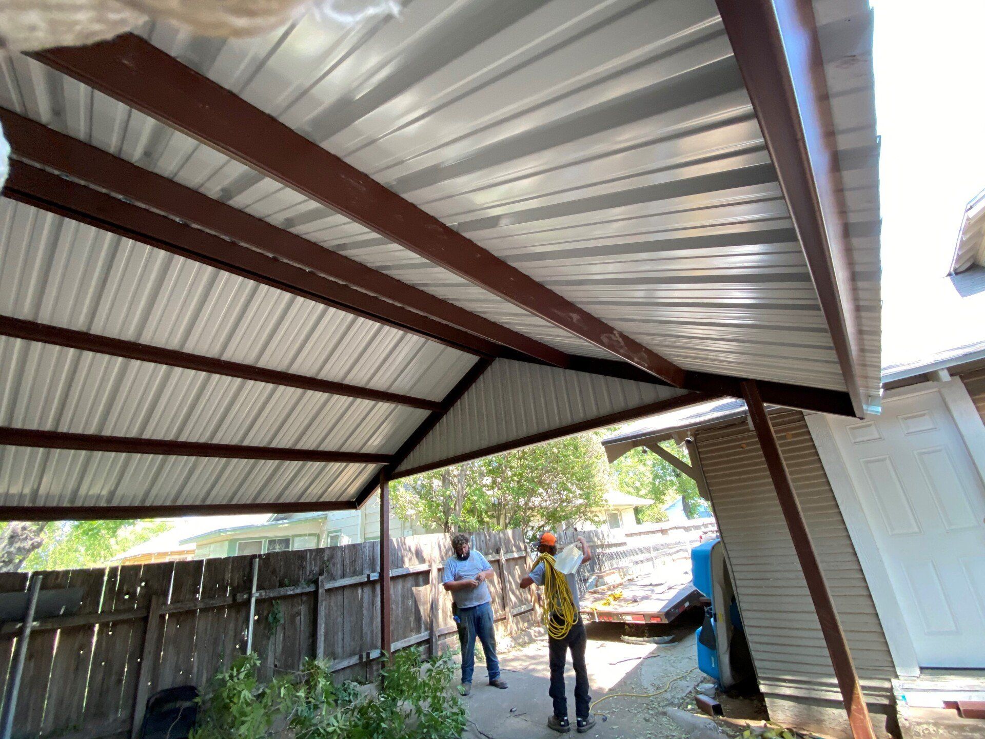 Two men are standing under a metal roof in front of a house.