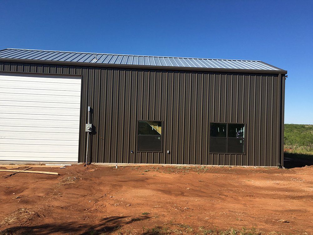 A large metal building with a white garage door is sitting in the middle of a dirt field.