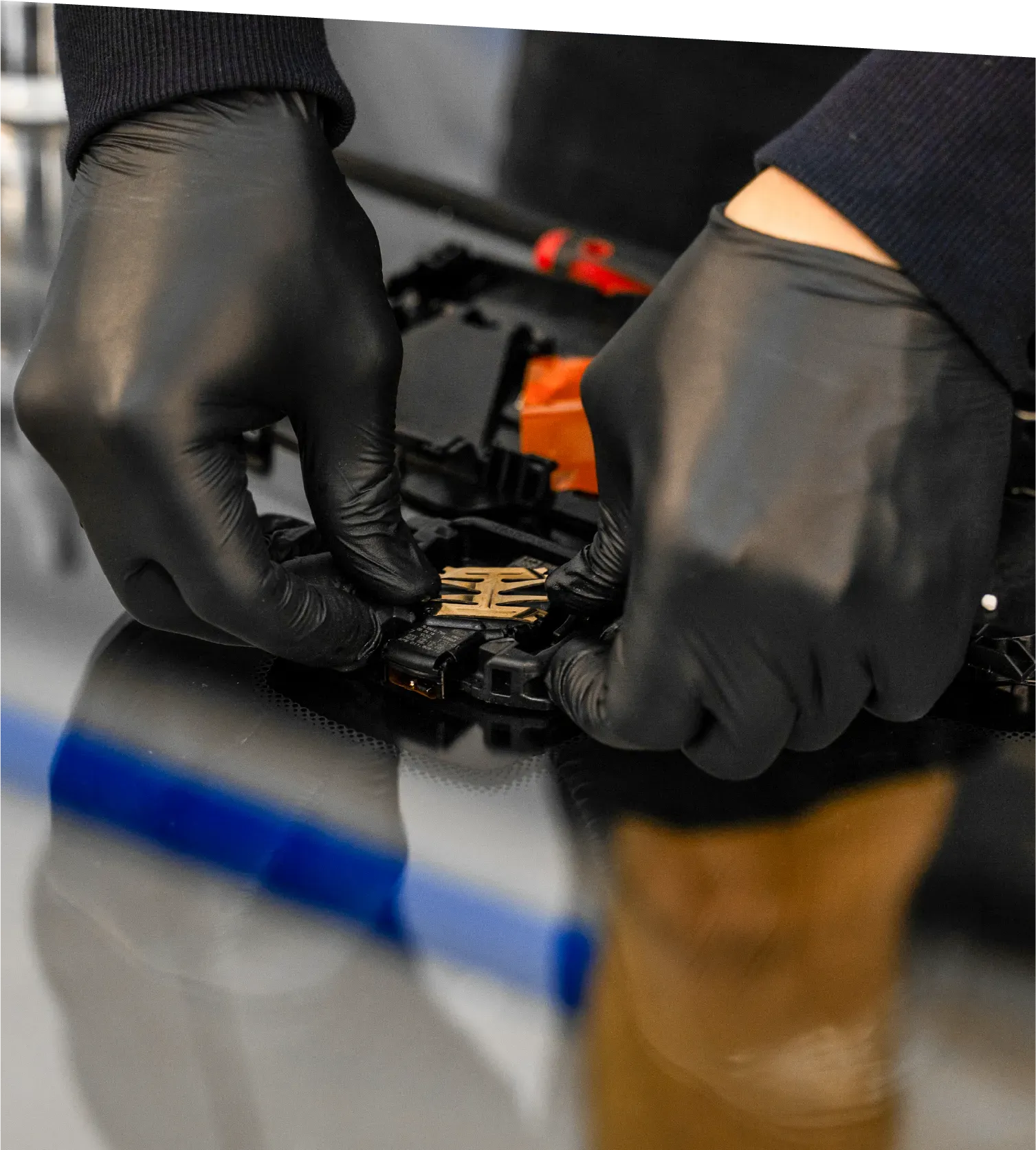 Gloved hand holding an orange and black multimeter with red and black probes, in front of a gray surface.
