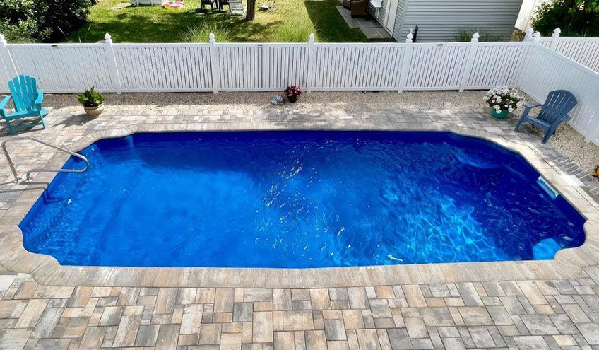 Blue swimming pool surrounded by a brick patio and white fence, with two Adirondack chairs.