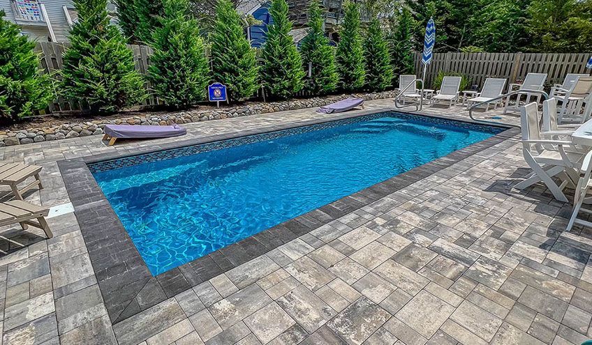 Rectangular blue swimming pool surrounded by gray pavers and lounge chairs. Trees line the back fence.