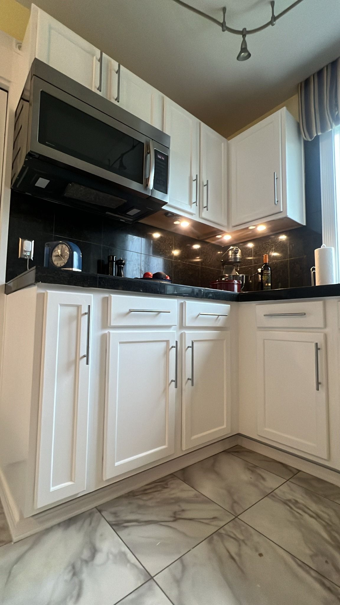 White kitchen cabinets with a black countertop, stainless steel microwave, and marble floors.