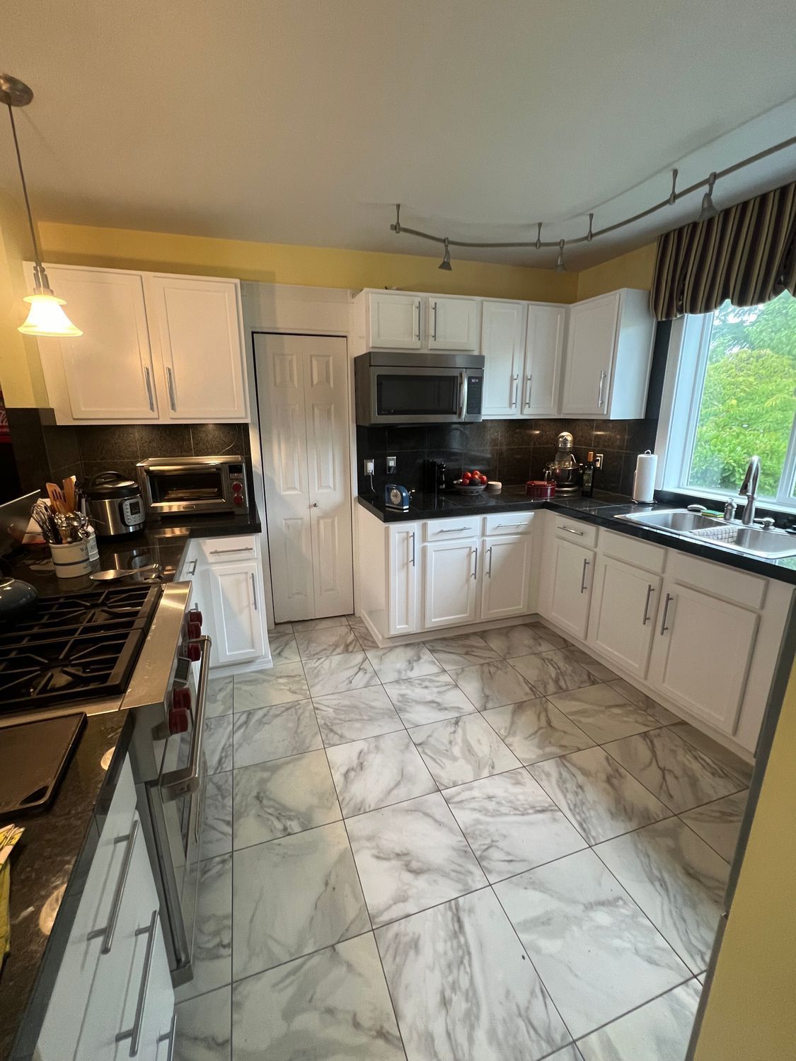 White kitchen with marble-look tile floor, white cabinets, black countertops, and a window with a patterned curtain.