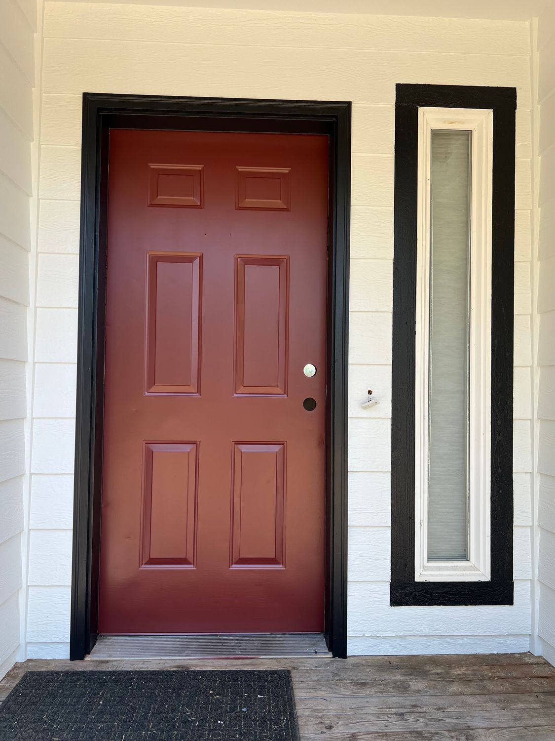 Red door with black trim and a tall window on a white wall. A doormat is in front of the door.