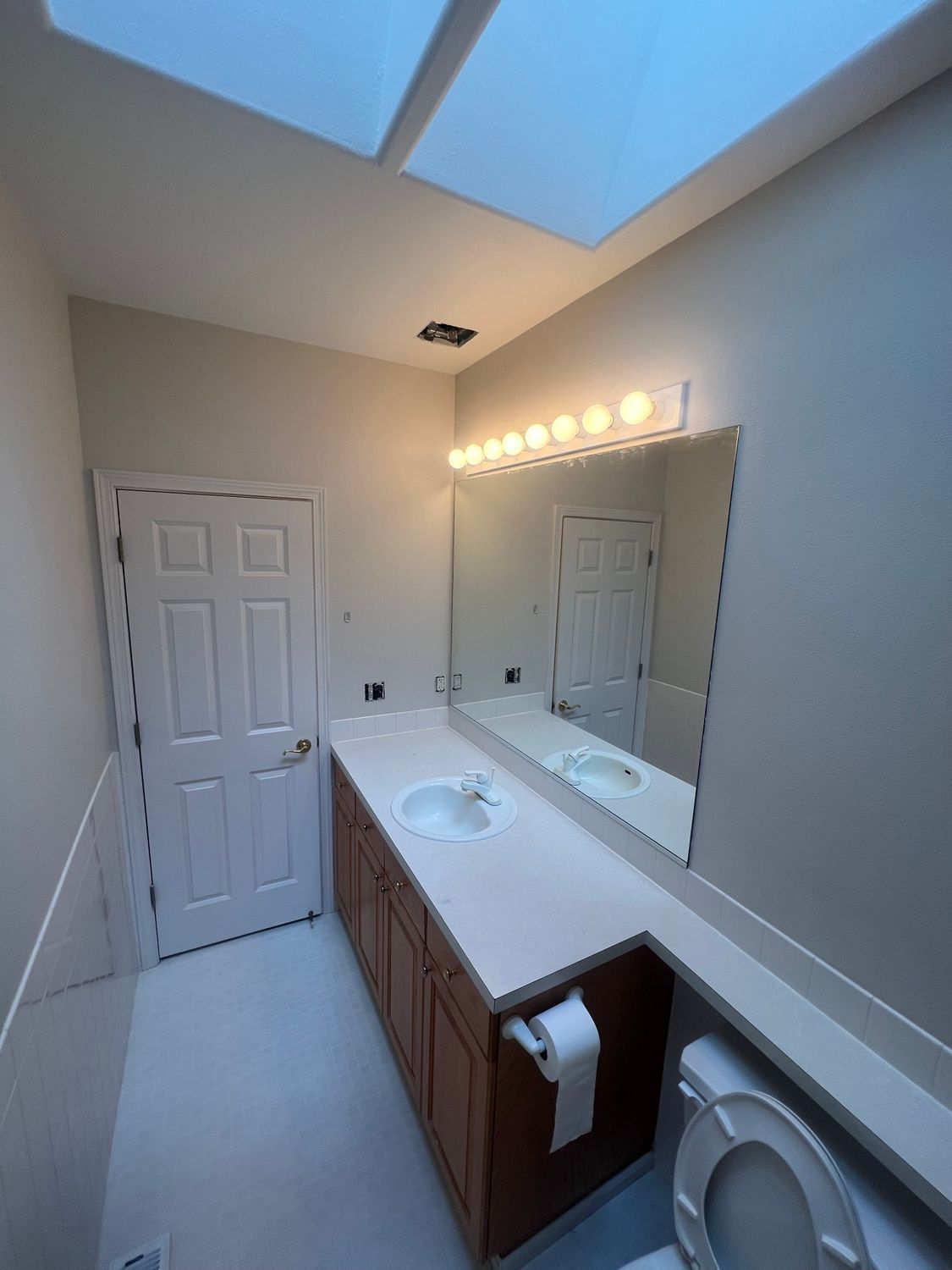 Bathroom with vanity, mirror, toilet, and door, lit by overhead light and skylights.