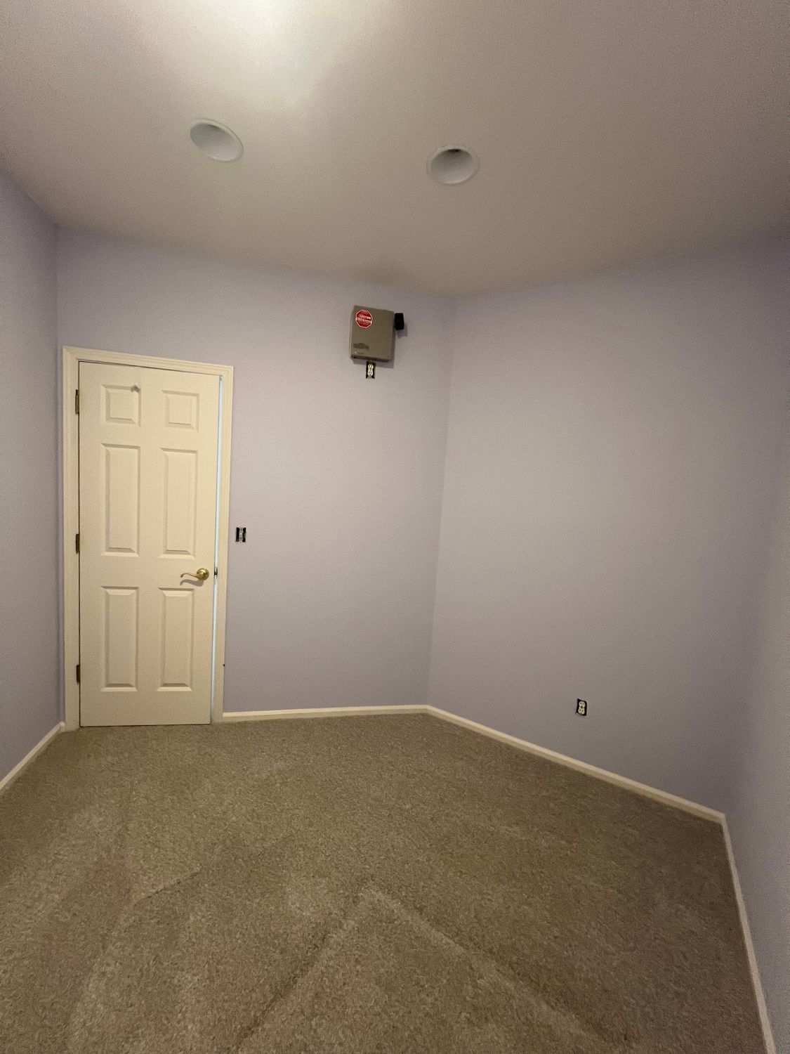 Empty room with lavender walls, white door, and beige carpet. Recessed lights and a fuse box on the ceiling.