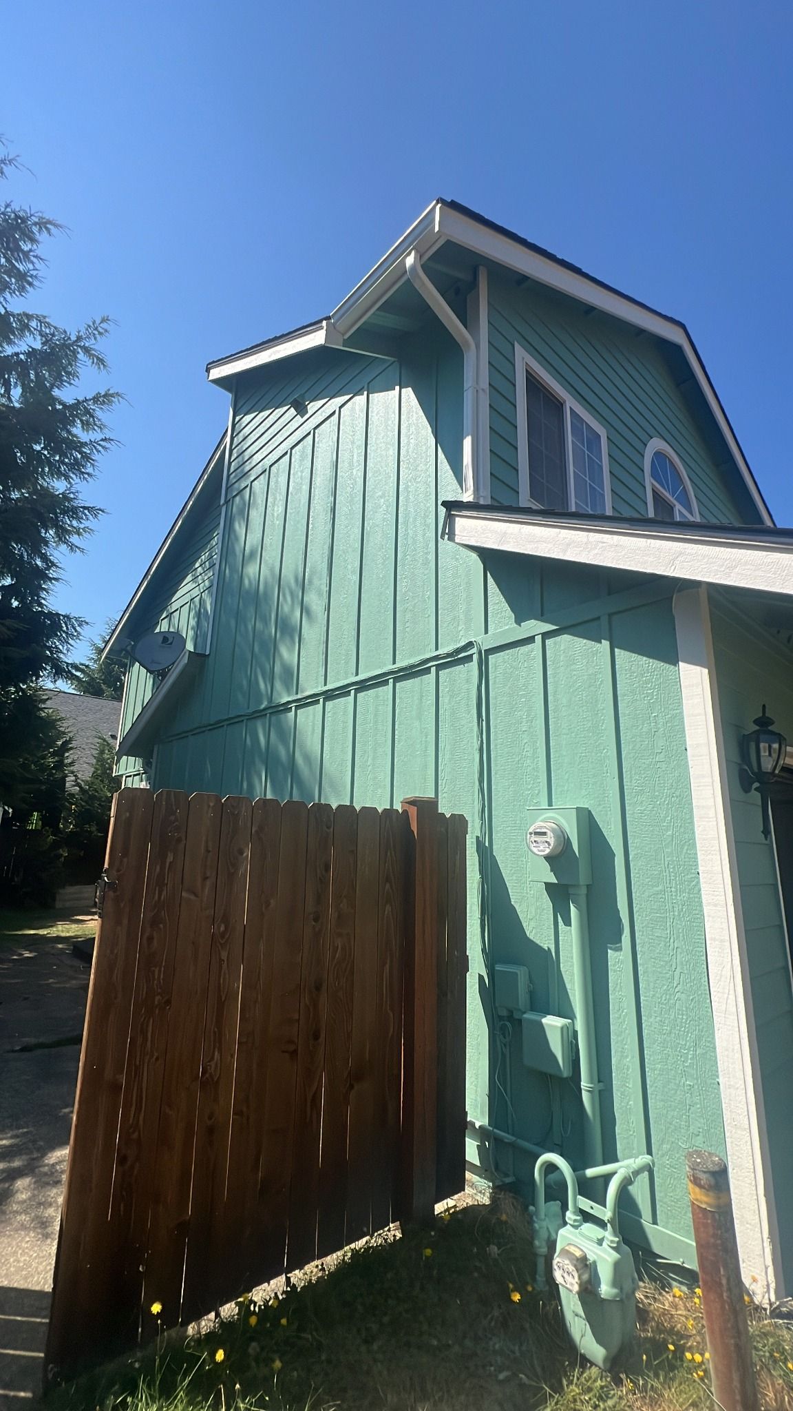 Green house with white trim and gabled roof, wooden fence, gas meter, and sunny blue sky.