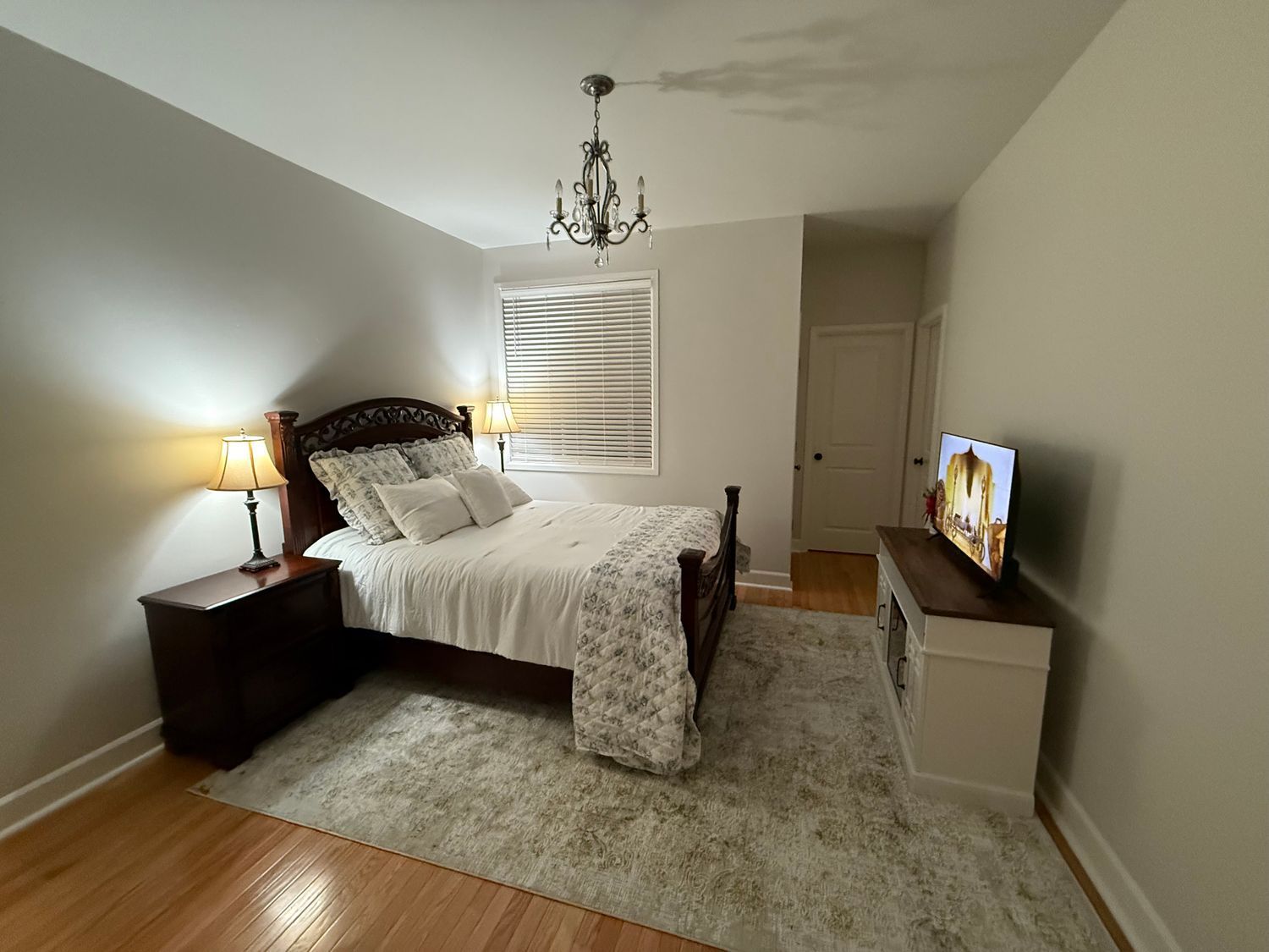 Bedroom with a bed, nightstands, rug, TV, and chandelier. Light walls and wood floor.