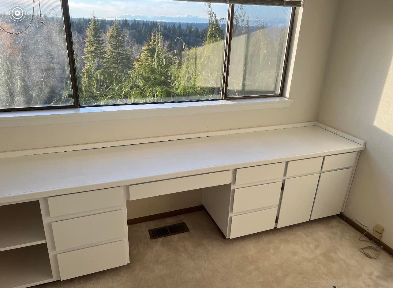 Built-in white desk with drawers under a large window with a forest view.