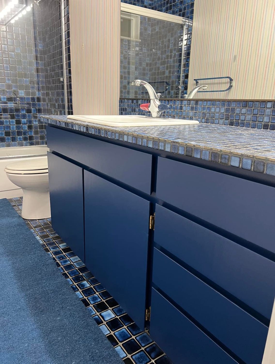 Blue bathroom with mosaic tile and vanity, white sink, toilet, and rug.