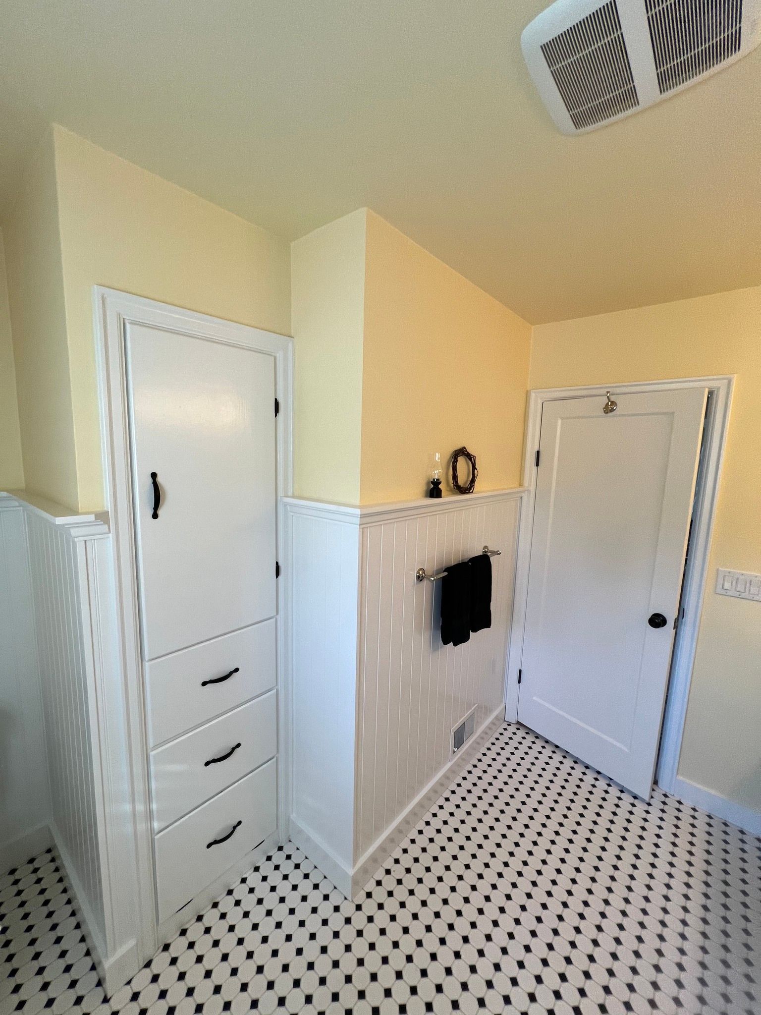 Bathroom with black and white tiled floor, white and yellow walls, tall white storage cabinet.