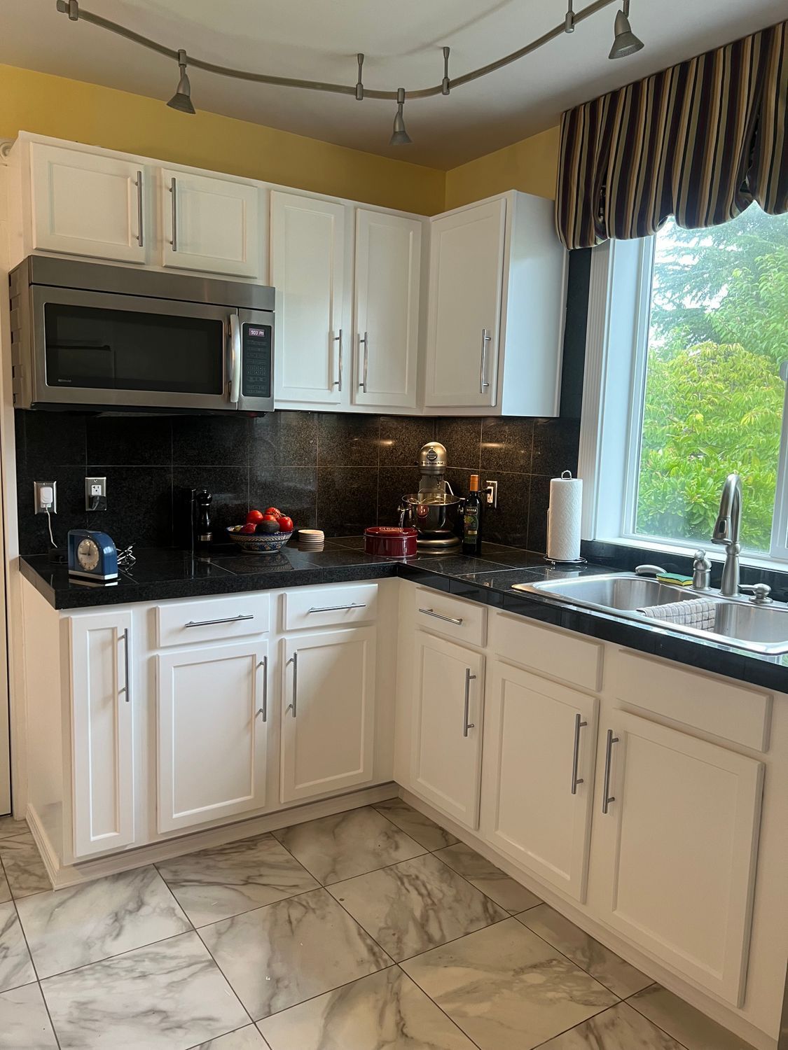 White kitchen cabinets, black countertops, stainless steel appliances. Corner window with view of greenery.