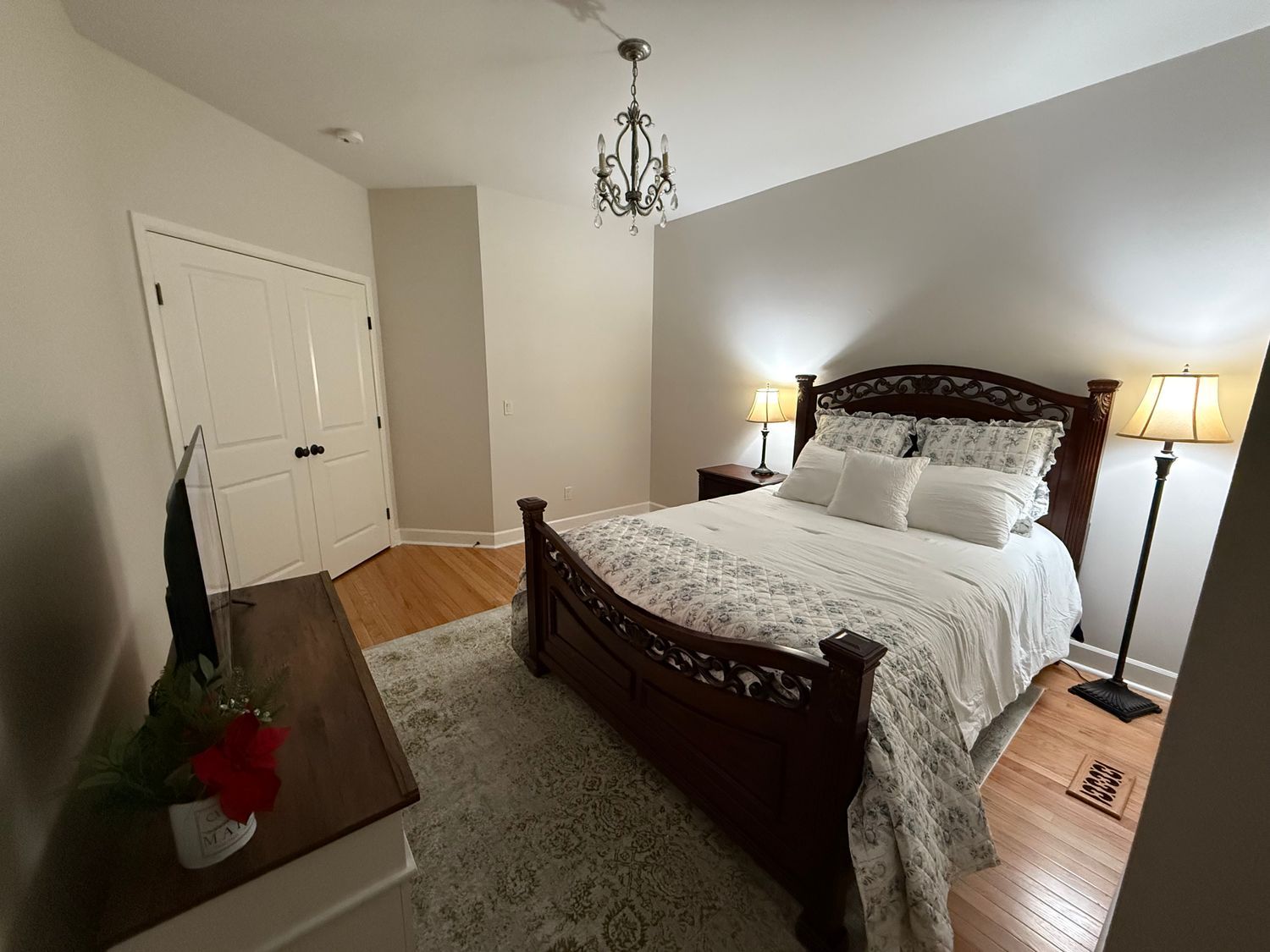 Bedroom with a wooden bed, dresser, and chandelier. Soft lighting, neutral walls, and rug.